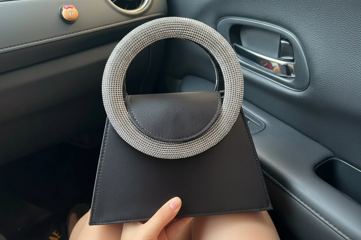 A black handbag with a large circular handle encrusted with silver rhinestones, held by a person sitting inside a car.
