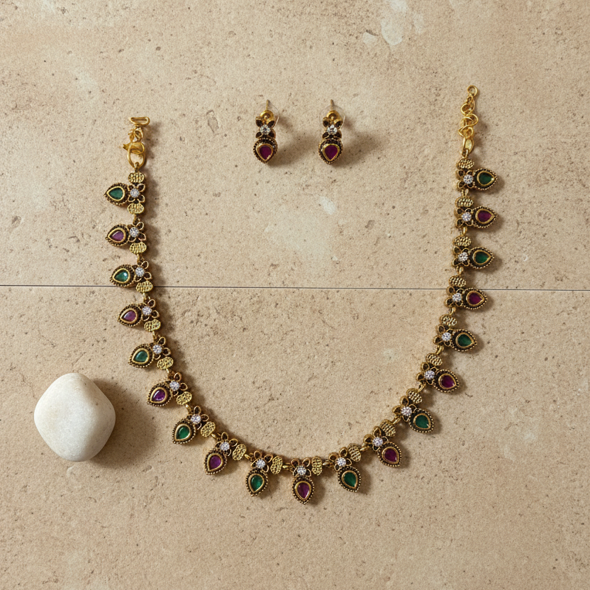 Gold-tone jewelry set featuring a necklace and matching earrings with small red and green teardrop-shaped stones, displayed on a white fabric jewelry bust.