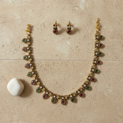 Gold-tone jewelry set featuring a necklace and matching earrings with small red and green teardrop-shaped stones, displayed on a white fabric jewelry bust.