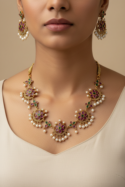 An ornate gold-toned jewelry set displayed on a white fabric bust, featuring a necklace and matching earrings with floral motifs, pearl-like beads, and red and green stone accents.