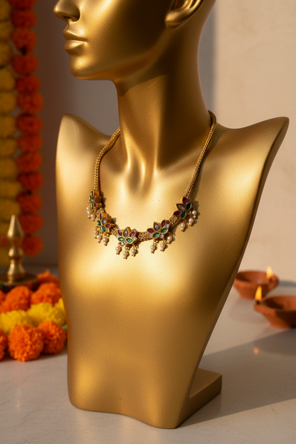 Gold-toned traditional necklace and matching flower-shaped earrings displayed on a white jewelry bust. The necklace has floral motifs with green and red enamel details and small pearl-like drops, with a long tassel hanging from one side.