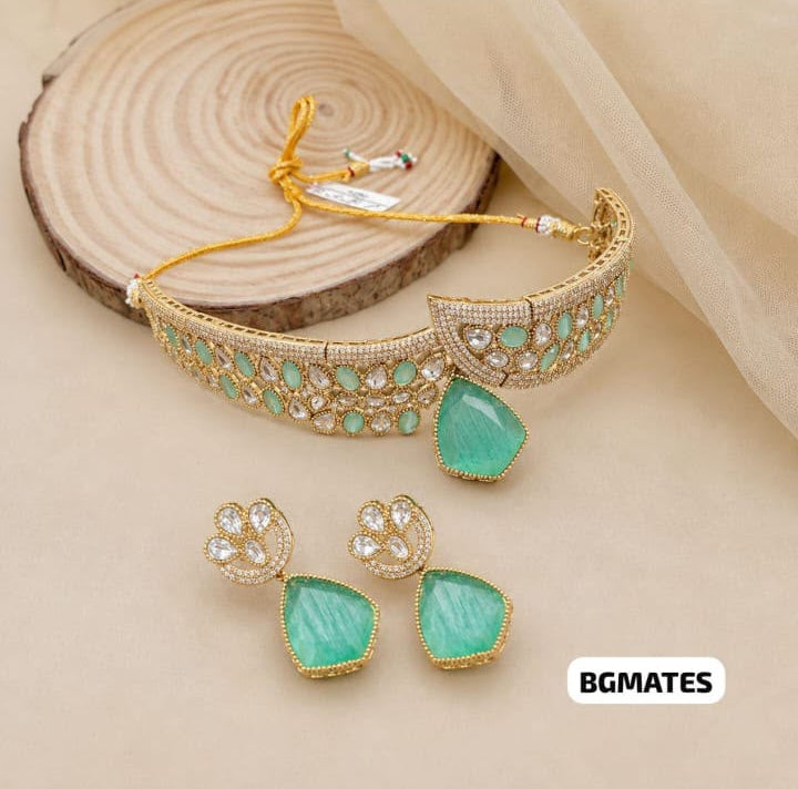 Gold-toned jewelry set featuring a curved bracelet with green stone accents, matching stud earrings with green teardrop-shaped stones, and a small gold clasp, displayed on a beige surface with a wood slice prop.