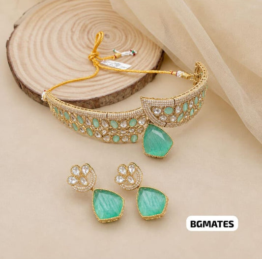 Gold-toned jewelry set featuring a curved bracelet with green stone accents, matching stud earrings with green teardrop-shaped stones, and a small gold clasp, displayed on a beige surface with a wood slice prop.