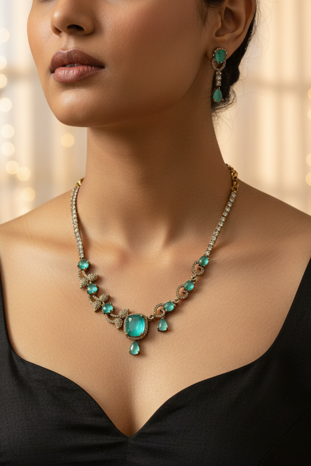 An ornate gold-toned necklace and matching drop earring set displayed on a cream fabric jewelry bust. The jewelry features prominent green teardrop-shaped stones and smaller clear stones set in a textured gold-tone metal.