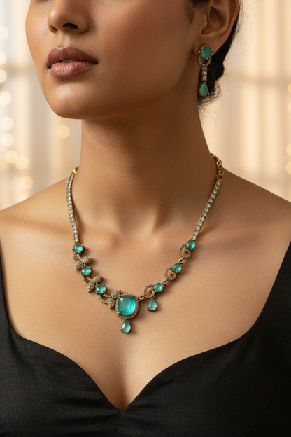 An ornate gold-toned necklace and matching drop earring set displayed on a cream fabric jewelry bust. The jewelry features prominent green teardrop-shaped stones and smaller clear stones set in a textured gold-tone metal.