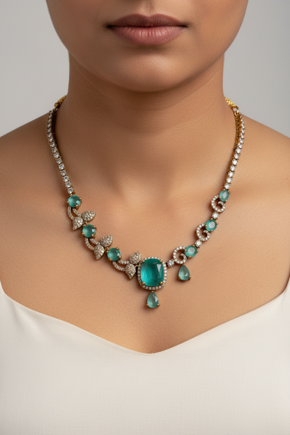 An ornate gold-toned necklace and matching drop earring set displayed on a cream fabric jewelry bust. The jewelry features prominent green teardrop-shaped stones and smaller clear stones set in a textured gold-tone metal.