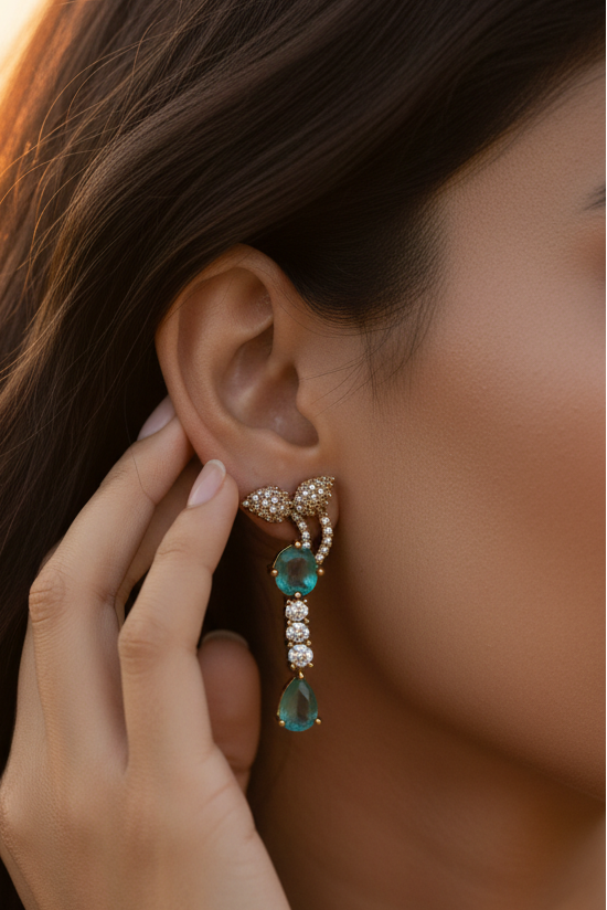 An ornate gold-toned necklace and matching drop earring set displayed on a cream fabric jewelry bust. The jewelry features prominent green teardrop-shaped stones and smaller clear stones set in a textured gold-tone metal.