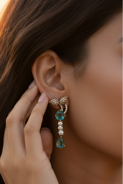 An ornate gold-toned necklace and matching drop earring set displayed on a cream fabric jewelry bust. The jewelry features prominent green teardrop-shaped stones and smaller clear stones set in a textured gold-tone metal.