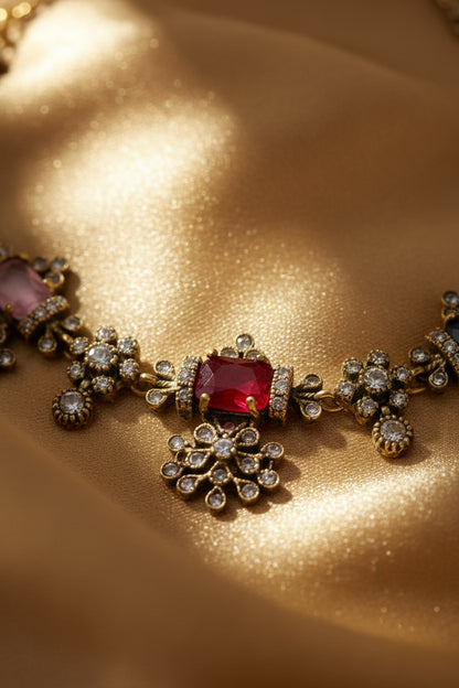A gold-toned traditional Indian jewelry set displayed on a white fabric jewelry bust. The set includes a necklace and a matching top piece, both decorated with small gold-toned floral motifs and colorful gemstones in green, red, pink, and purple.