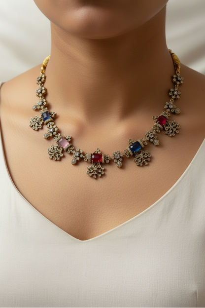 A gold-toned traditional Indian jewelry set displayed on a white fabric jewelry bust. The set includes a necklace and a matching top piece, both decorated with small gold-toned floral motifs and colorful gemstones in green, red, pink, and purple.