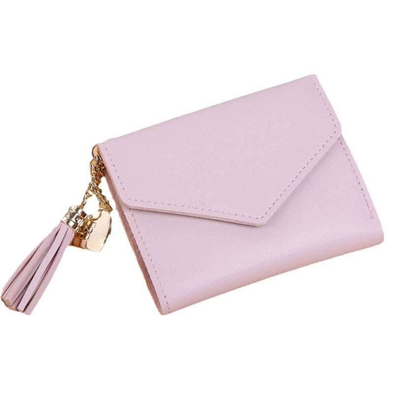 A light pink faux leather wallet with an envelope-style flap and a gold-tone tassel charm, shown on a plain white background.